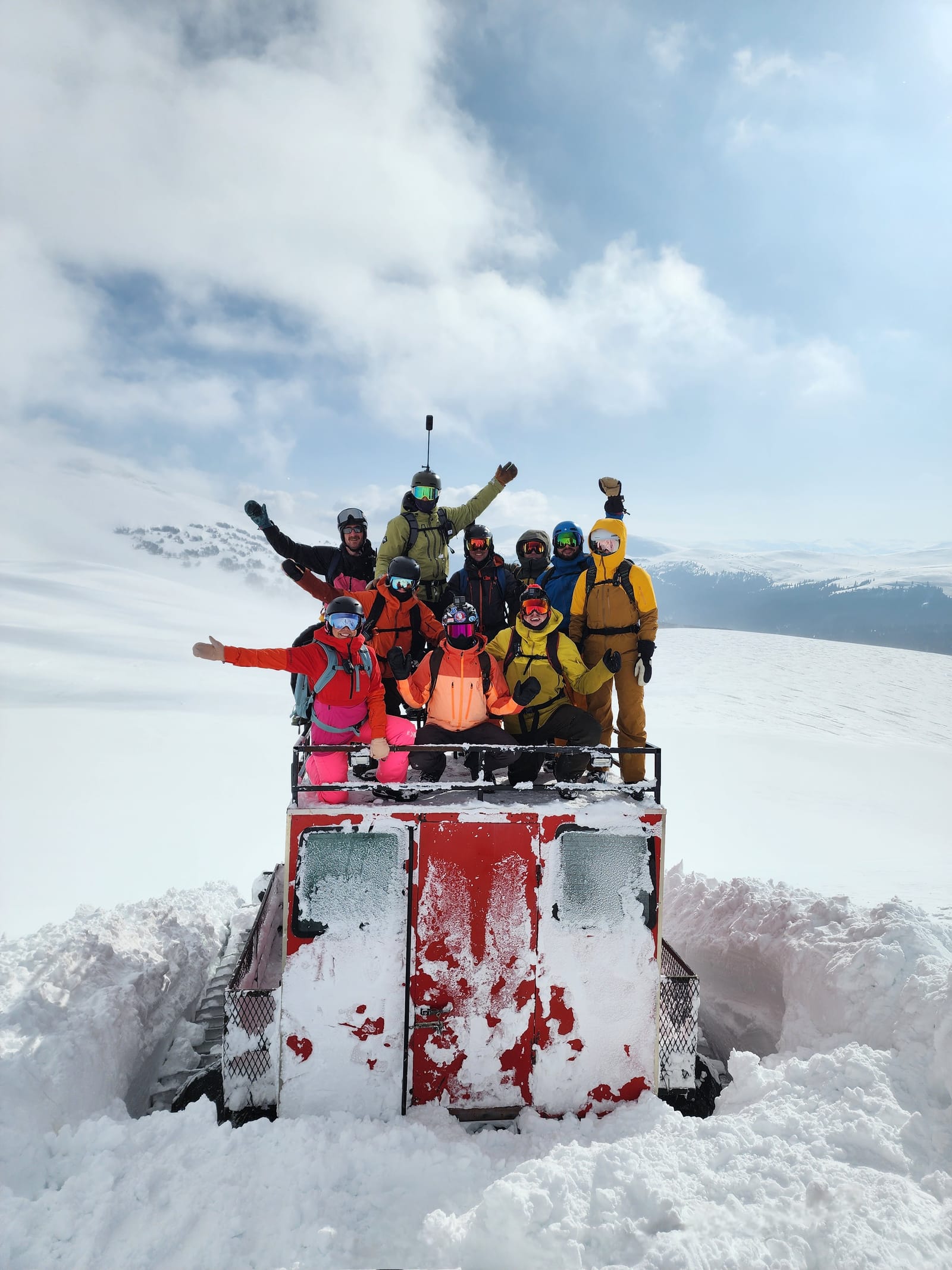 About ten riders waving from the roof of a red snowcat on a snowy mountain