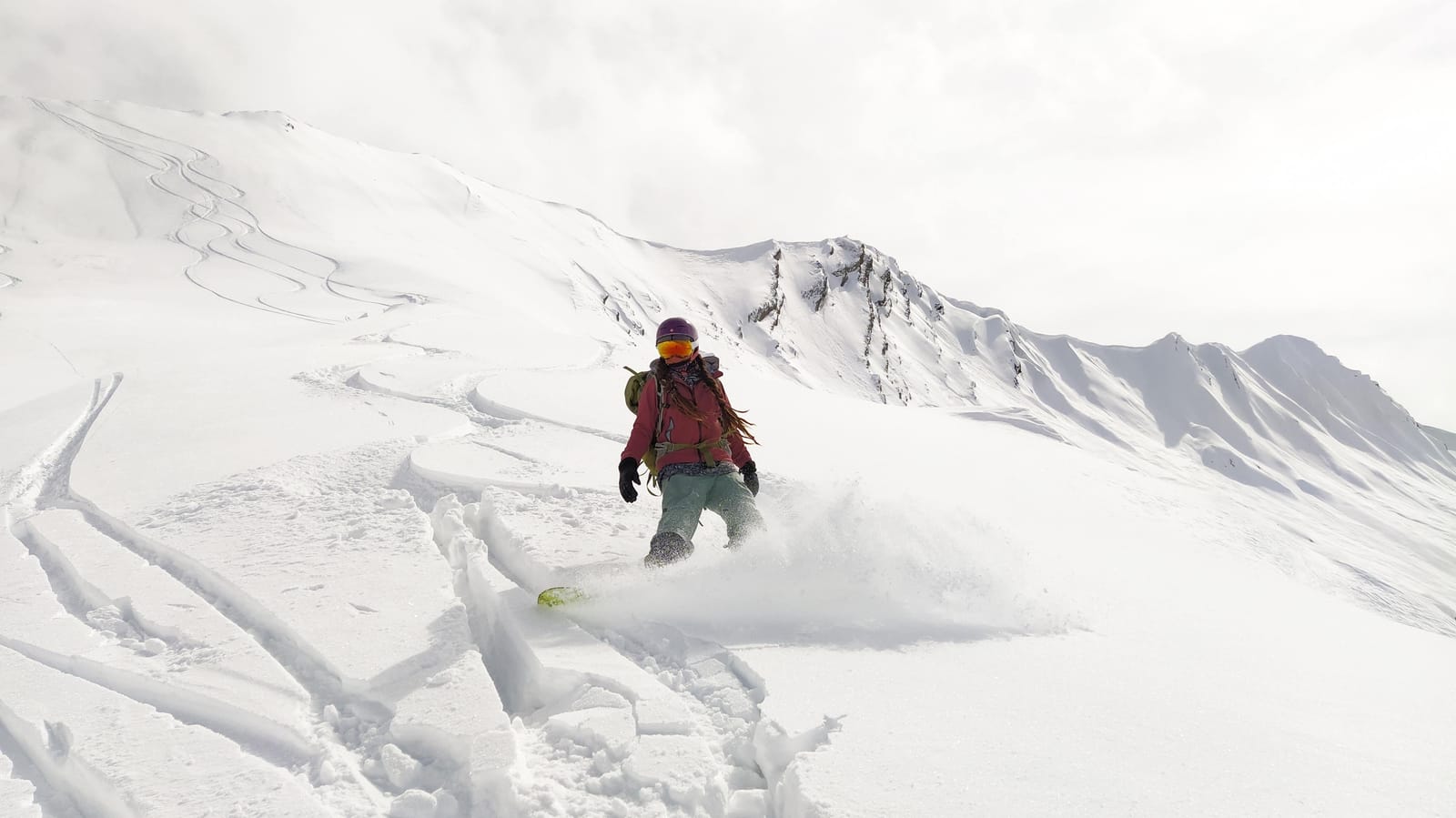 A snowboarder in a red jacket carving through fresh powder below a snowy ridge in Gudauri