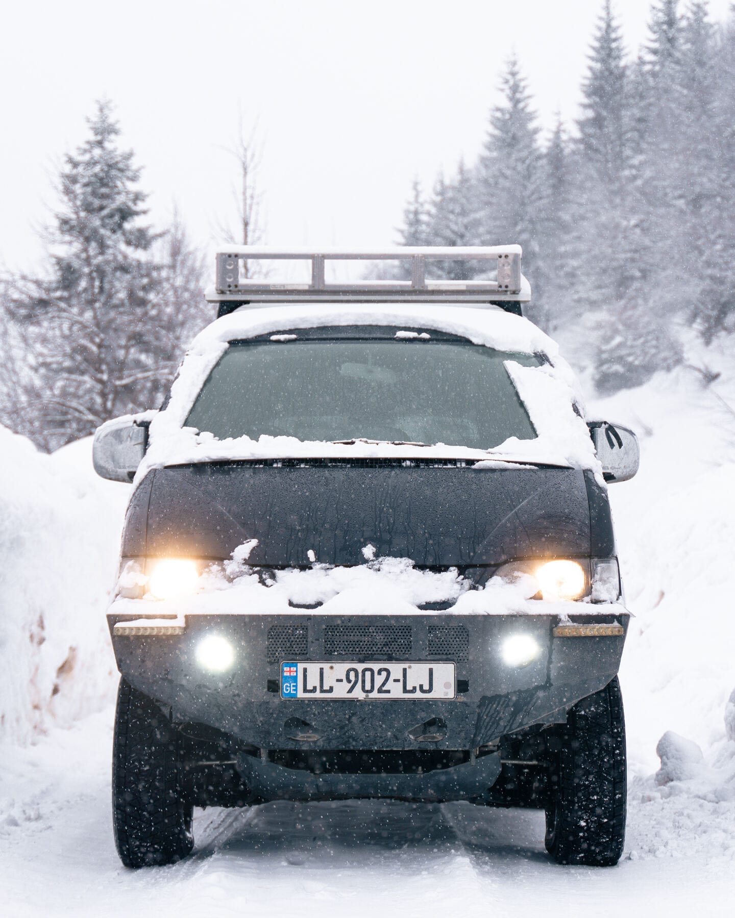 A snow-covered 4x4 van with its headlights on, driving down a snowy forest road in falling snow